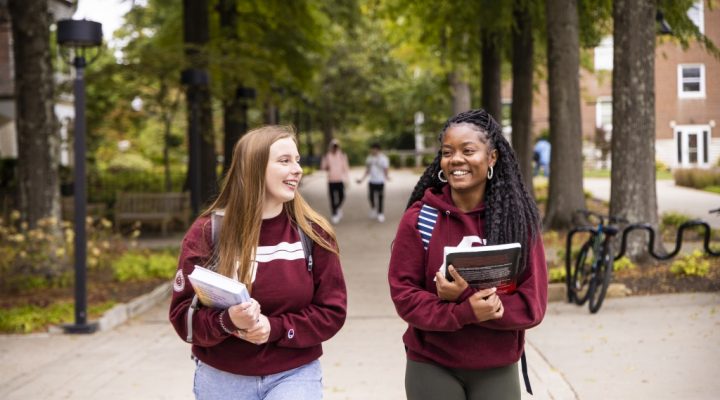Two students walk and talk on the sidewalk while holding textbooks.