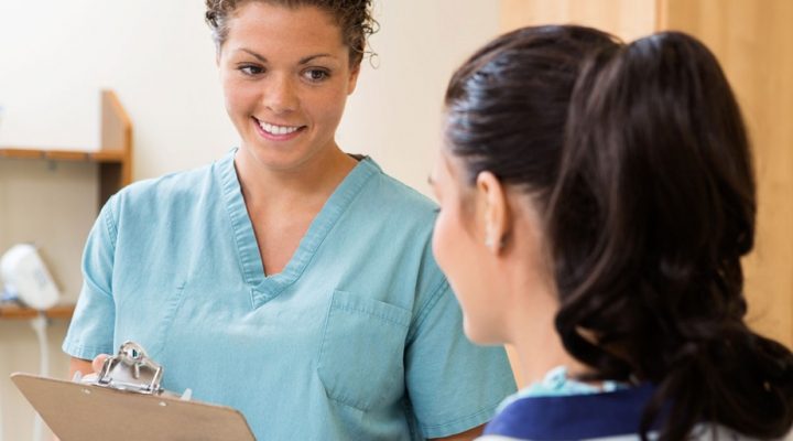An image of someone in scrubs holding a clipboard talking to a patient.