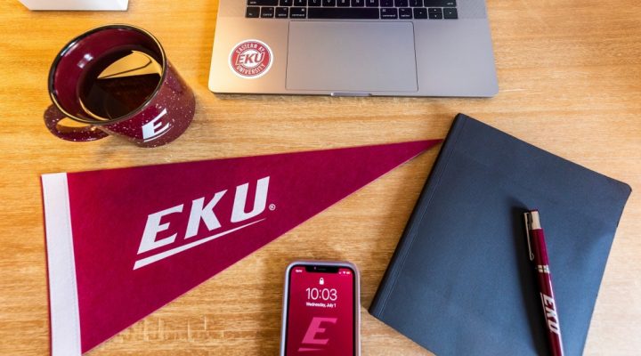 A laptop with an EKU sticker, an EKU mug, an EKU flag, a notebook with an EKU pen, and a phone with an EKU background.