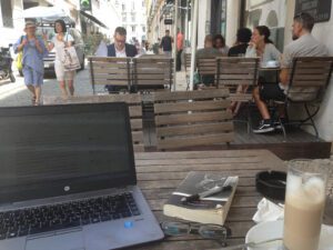 A laptop, coffe, and book on a table with people in the background.