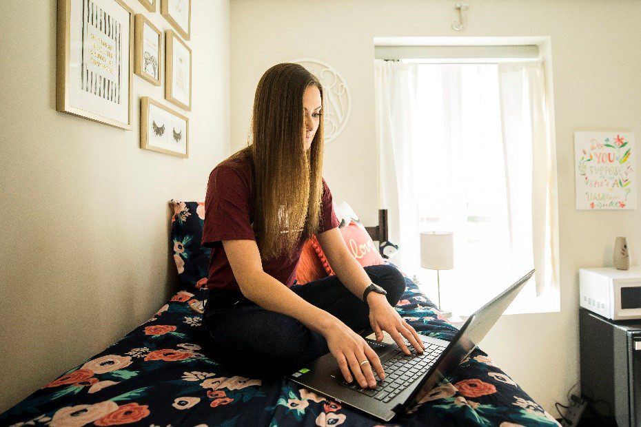 A student sitting on a bed in a dorm wearing an EKU shirt, while typing on the computer.