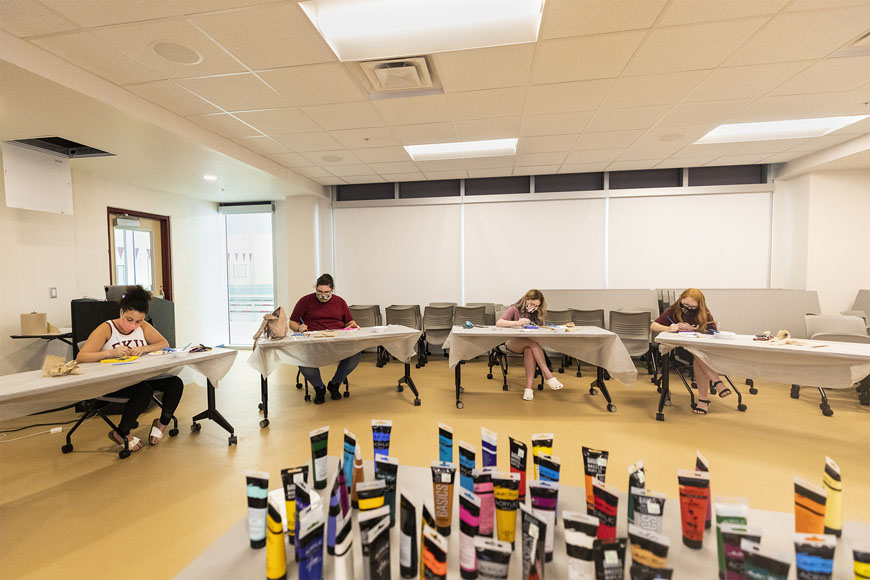 Four students sitting at tables spread apart painting canvases in the Rec Center