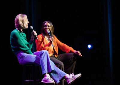 Two comedians seated on stage holding microphones laugh together