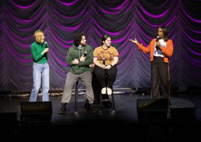 Two comedians speaking into microphones interview two audience members seated on stage