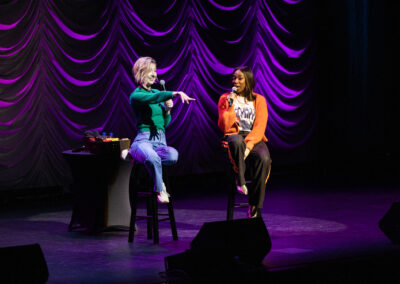 Two comedians seated on stage speaking into microphones, one points at the audience