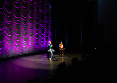 Two comedians in conversation on stage lit with a spotlight