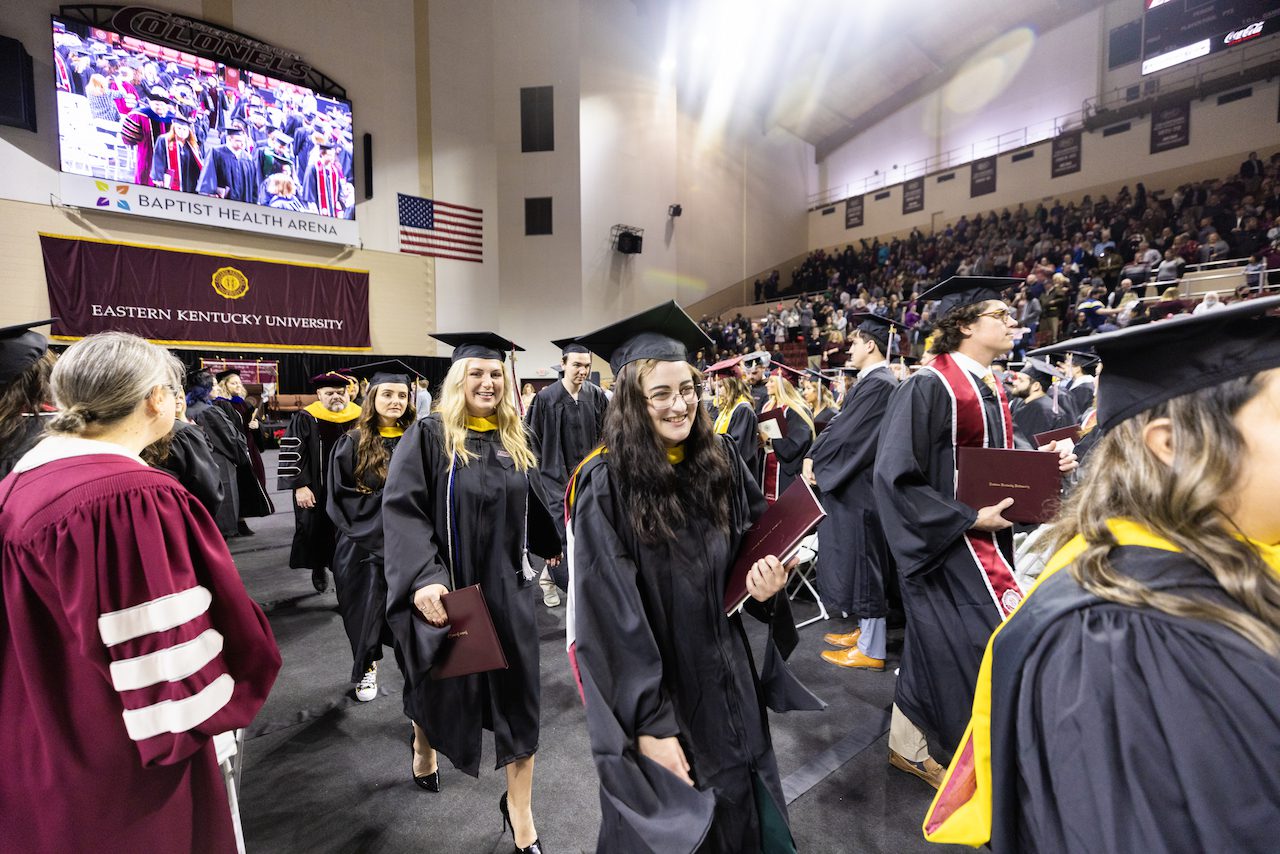 Smiling graduates with diplomas walking down aisle during commencement ceremony