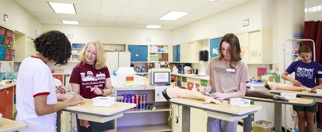 Three students in nursing setting learn from EKU instructor how to put in an IV into a practice arm.