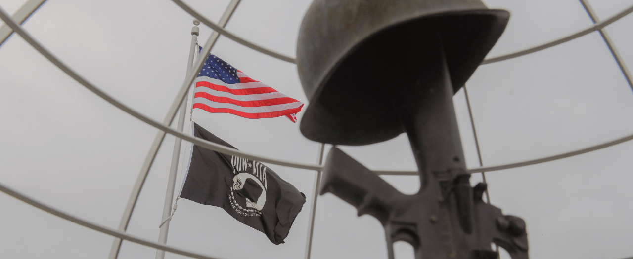 foreground closeup of sculpture of military gun with helmet and background flag pole with US flag and POW MIA flag