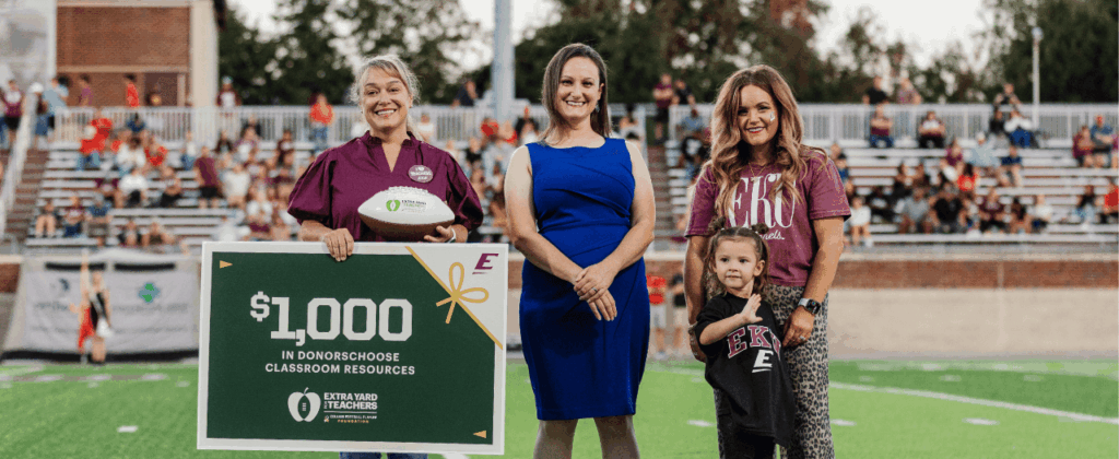 devon moody stands in a field with two other adults and a child with a sign from extra yard teachers showing that says $1,000 in donorschoose classroom resources