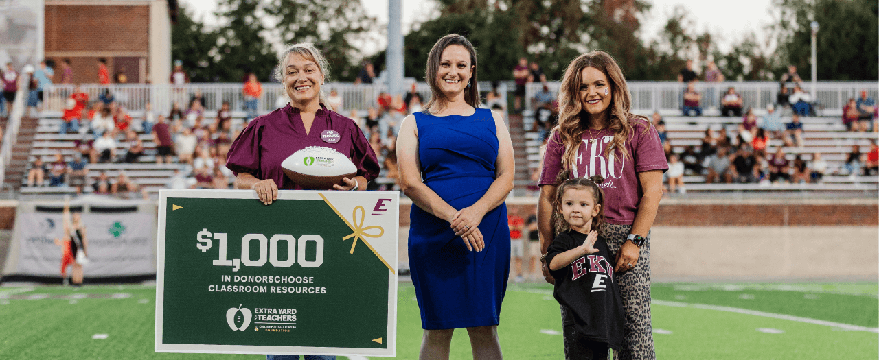 devon moody stands in a field with two other adults and a child with a sign from extra yard teachers showing that says $1,000 in donorschoose classroom resources