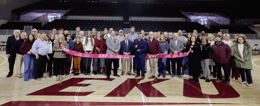 large group of people stand behind a ribbon in the floor of alumni coliseum