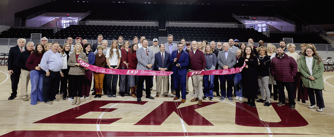large group of people stand behind a ribbon in the floor of alumni coliseum