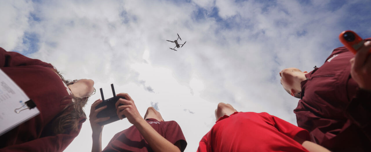 In a from-the-ground view, 4 students look at a drone in the sky as one of them pilots it with controls.
