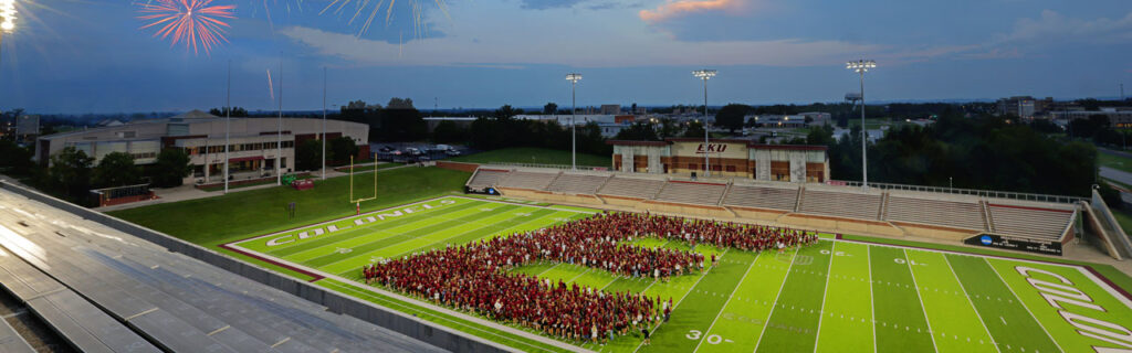 Students form the "big E" on the CG Bank Field Roy Kidd Stadium football field at night with fireworks in the background.