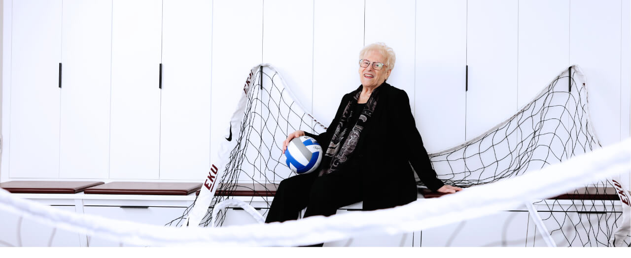 Banner image of Geri Polvino sitting with a ball on a volleyball net
