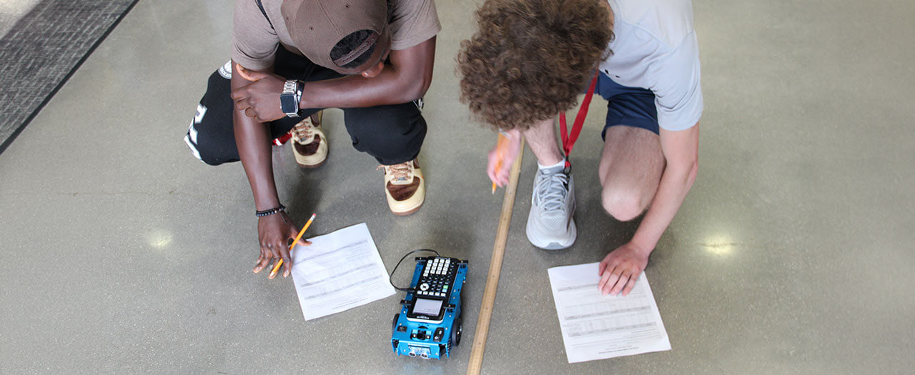 Two STEM Start students crouch to take notes about robotic car next to them